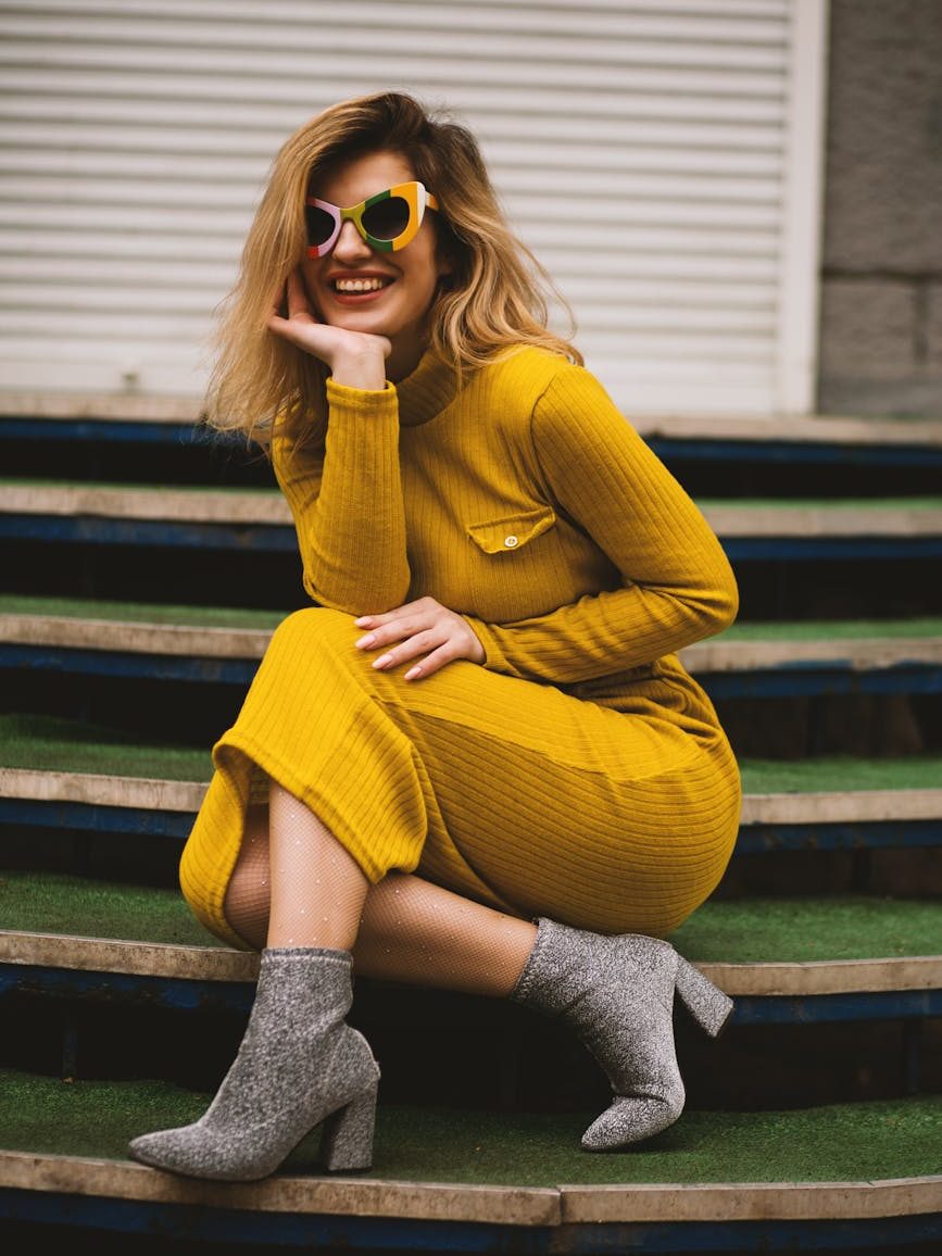 A woman with shoulder length blond hair sits on green stairs smiling. She wears large colourful plastic sunglasses and golden knit dress, sparkly hose and grey ankle boots.