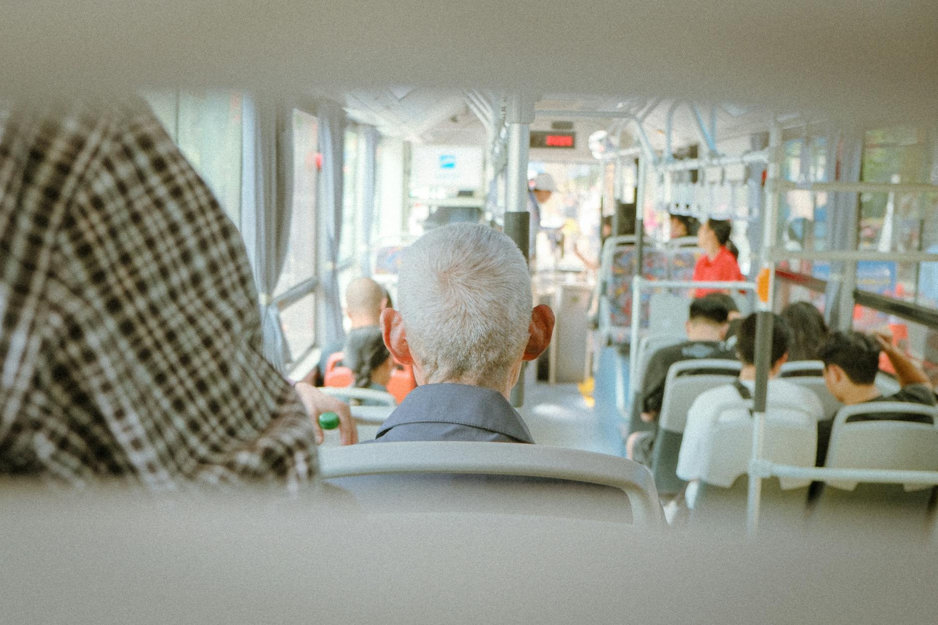 The back of an older man's grey head can be seen on a bus with others seated in front of him.