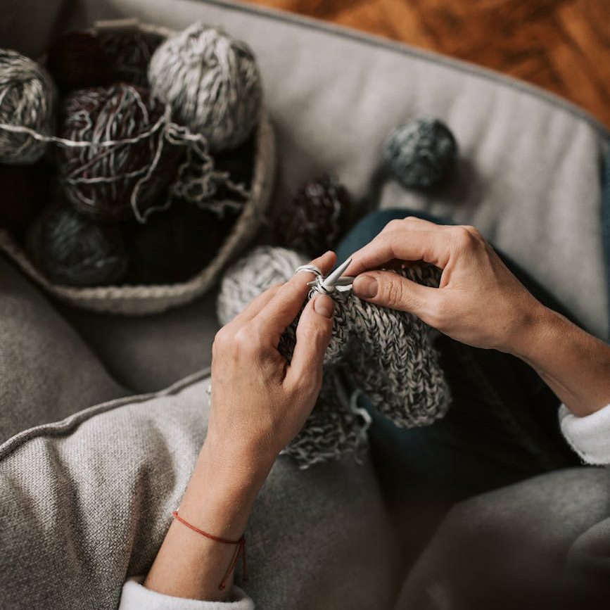 Hands holding knitting needles and knitting with grey yarn. A basket of various grey-coloured yarns is in the background.