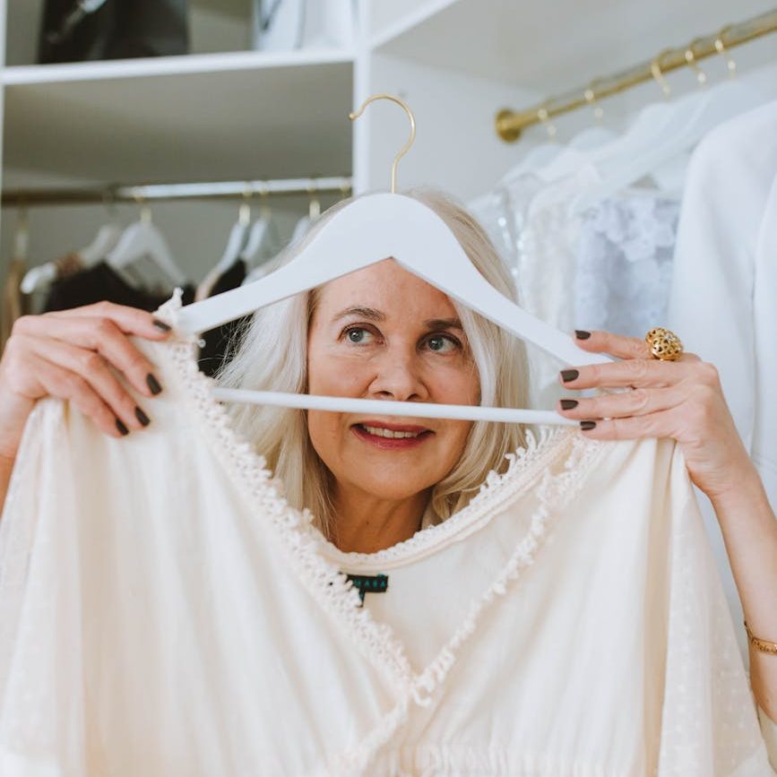 A woman holds a pale coloured blouse on a hanger in front of her. She appears to be deciding. 