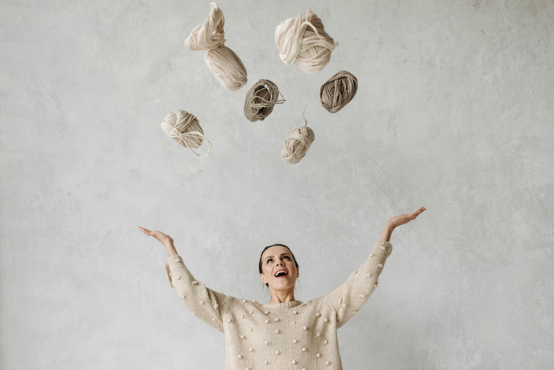 A woman in a beige sweater smiles while tossing beige and cream coloured yarns into the air.