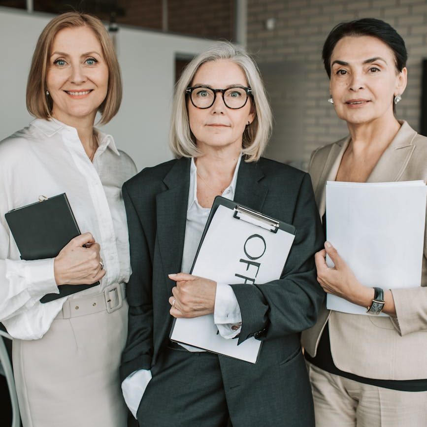 Three business women hold folders and papers.  The woman on the left wears a white shirt and cream skirt. The woman in the middle wears a grey pantsuit with white shirt. The woman on the right wears a cream pantsuit with a dark top underneath.