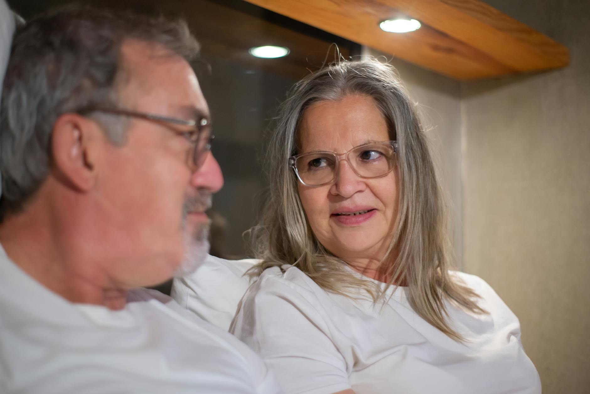 A woman with grey hair and glasses is turning to talk to a man with grey hair and glasses who is sitting beside her. 