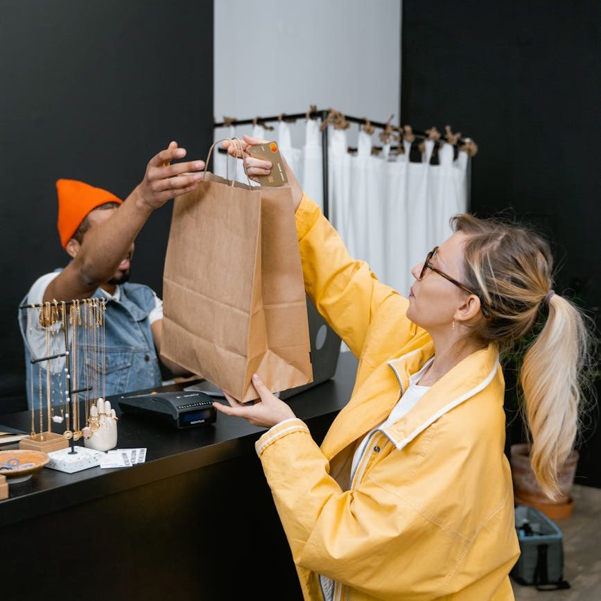 A woman in a yellow jacket is handed a brown shopping bag by the sales clerk on the other side of the counter. 