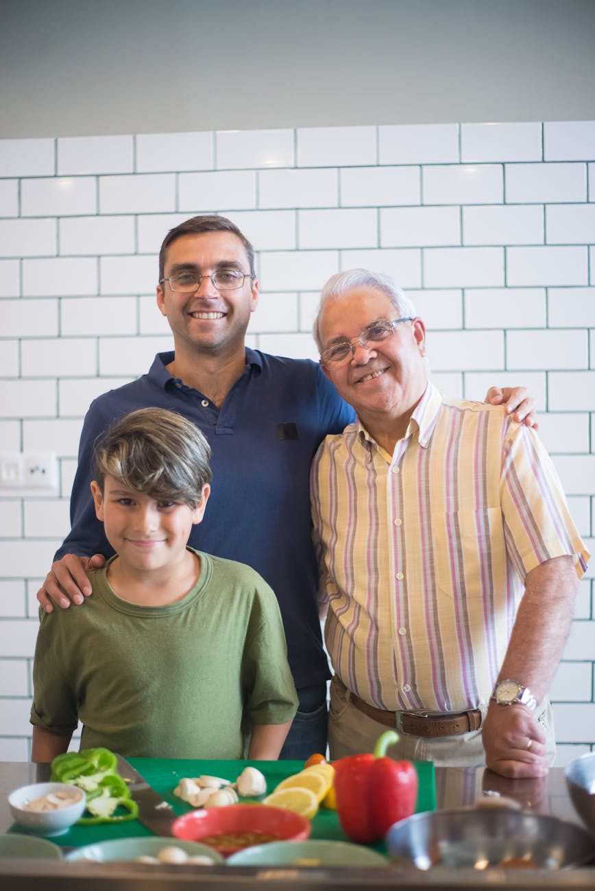 Three generations of males stand smiling behind a kitchen counter with vegetables on a chopping board in front of them.