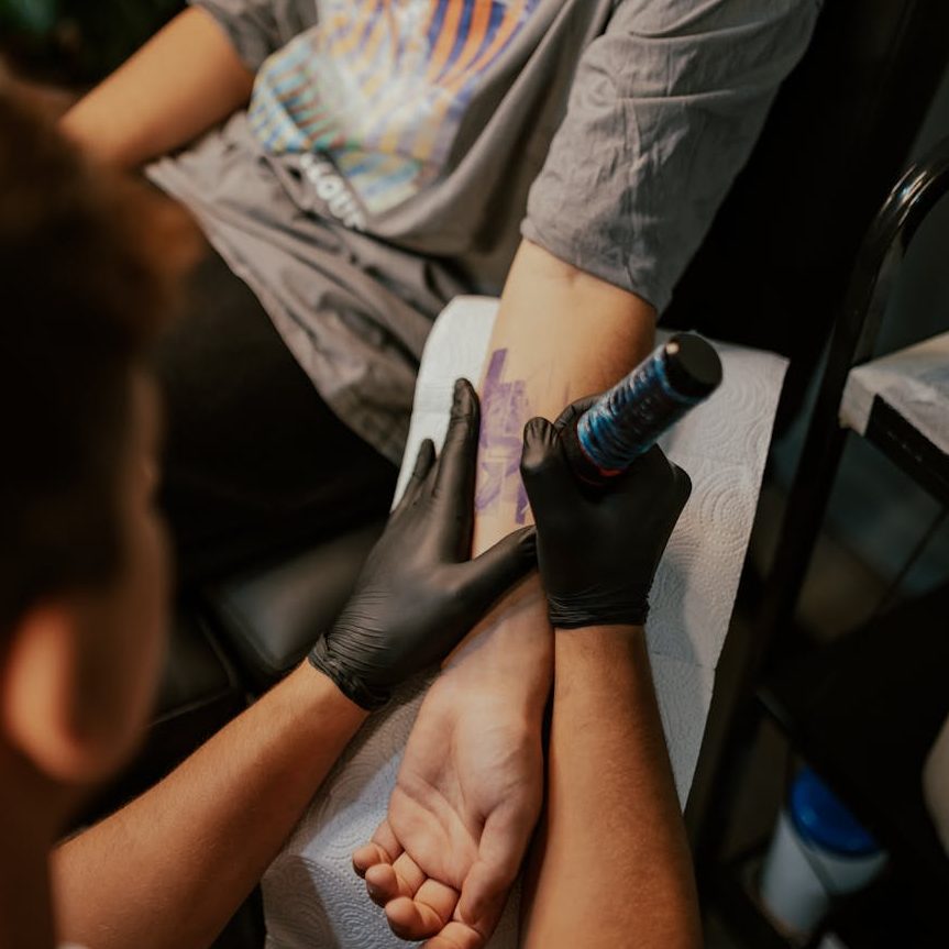 A person in a grey t-shirt getting their left inside forearm tattooed. The tattoo artist is wearing black gloves.