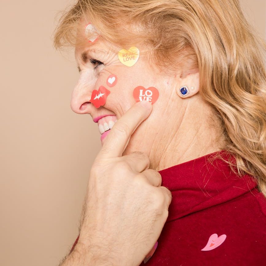 An older woman smiles with a variety of heart-shaped stickers on her face. A male hand points to the one that says, "Love."
