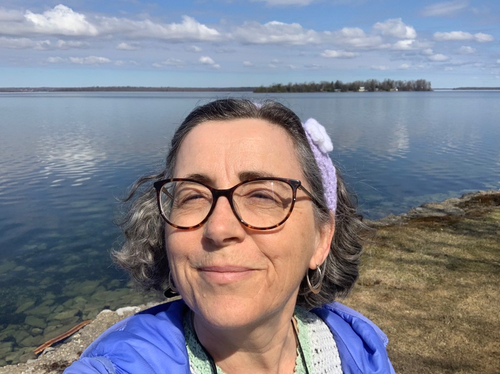 The author wearing a purple coat and hairband, smiling into the sunshine while standing in front of a calm lake.