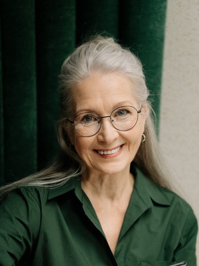 A woman with long grey hair and glasses, wearing a dark green blouse, is smiling at the camera.