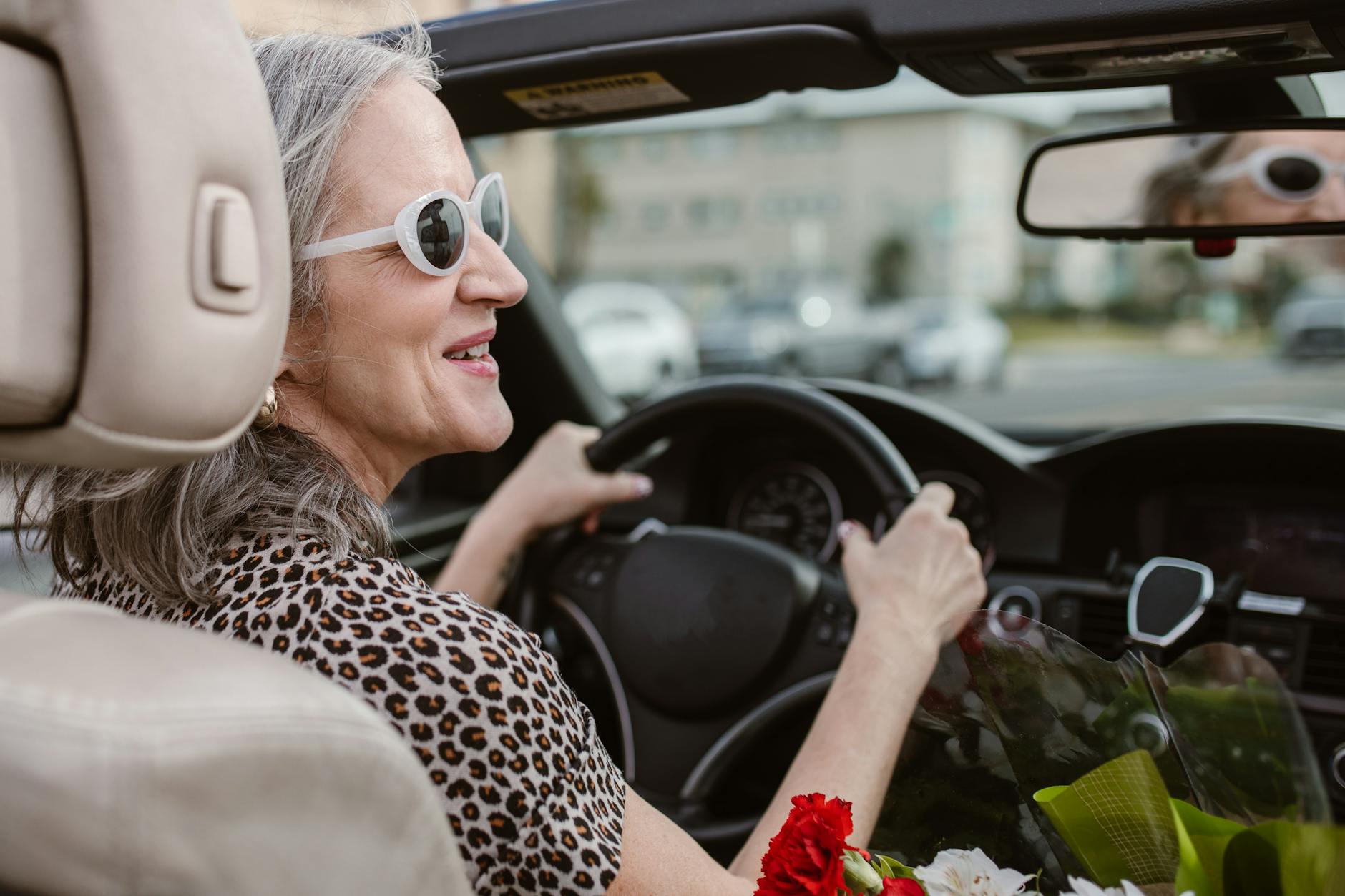 A woman with grey hair, white sunglasses and a leopard print top confidently drives a car.