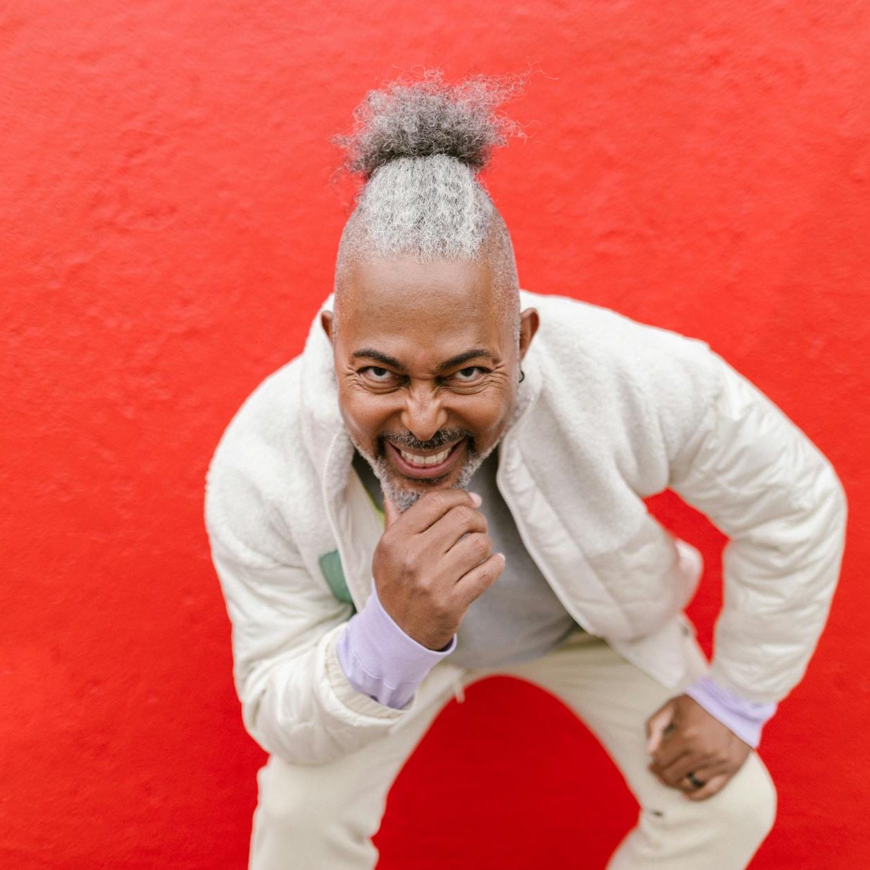 A man with in a light jacket and pants with grey hair tied back and a grey beard, leans smiling towards the camera.
