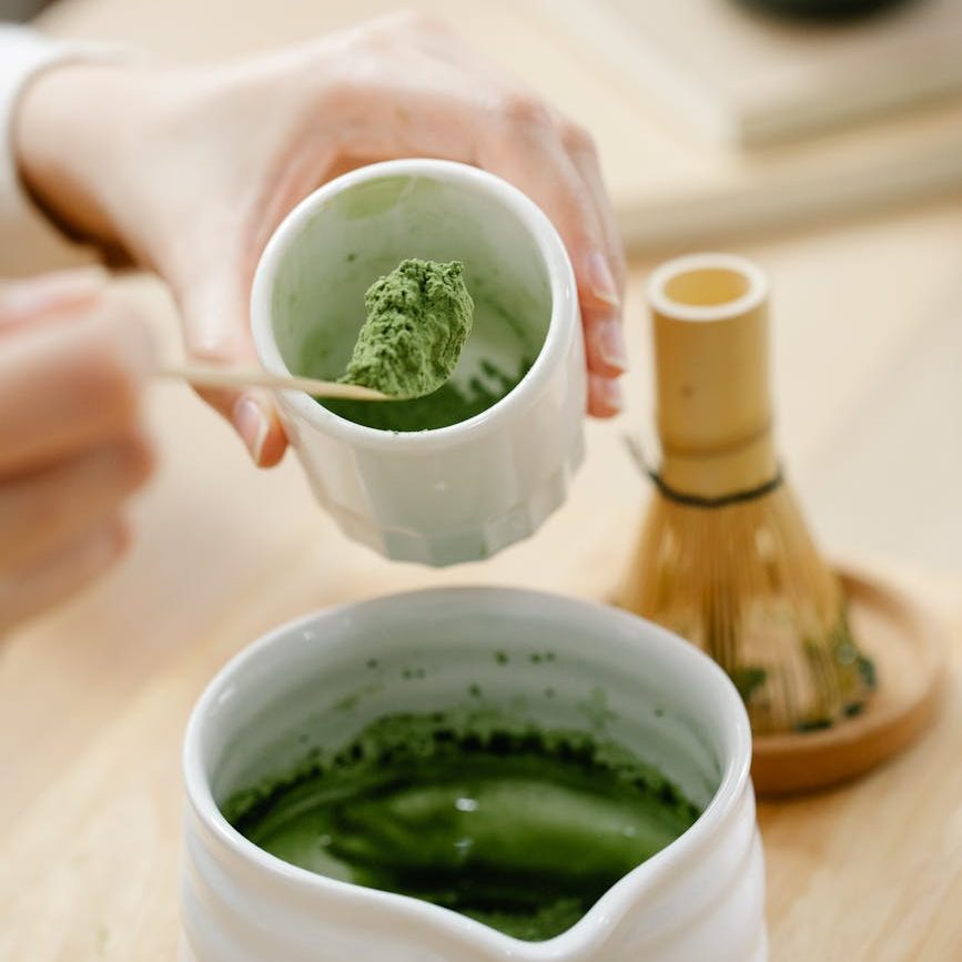 A person spoons matcha powder into a white jug. A bamboo matcha whisk sits in the background with green matcha paste on its base.