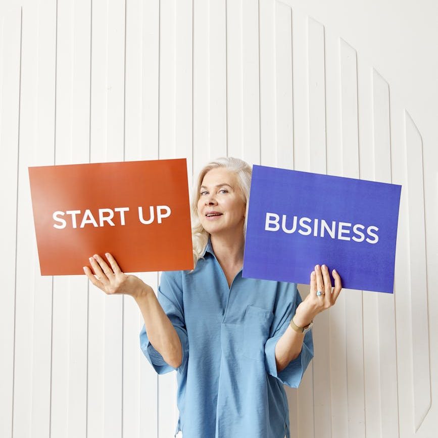 A grey haired woman holds a red sign that says, "Start up," and a blue sign that says, "Business."