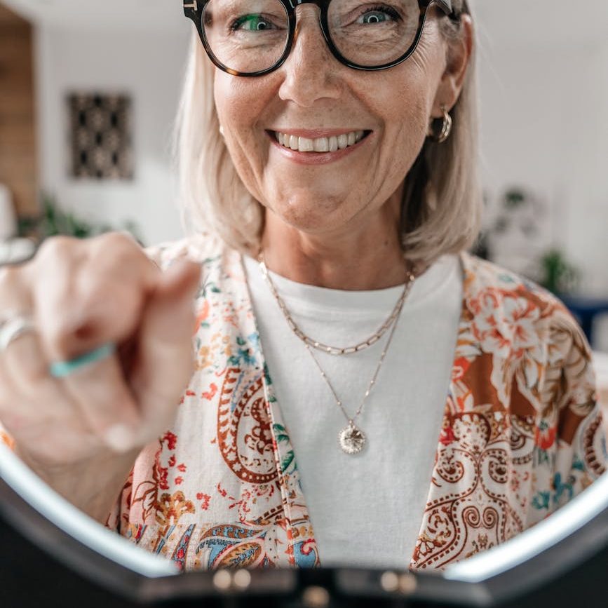 An older woman wearing glasses and  a printed top over a white t-shirt smiles as she points towards the camera.