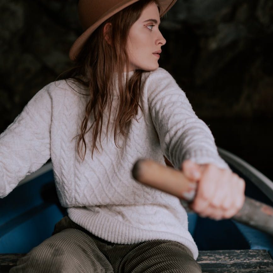 A woman in a brown hat, cream wool sweater and cord pants looks over her shoulder as she rows a boat.