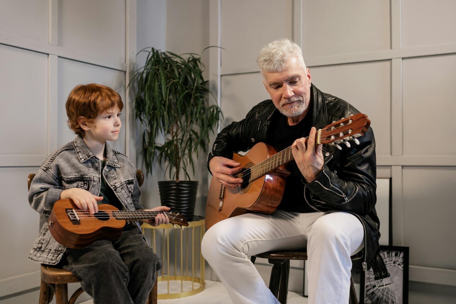 An older man playing guitar while a young boy holds a ukulele and watches him.