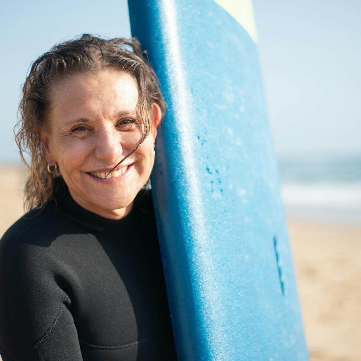A person in a wetsuit smiles broadly as she holds a blue surfboard.