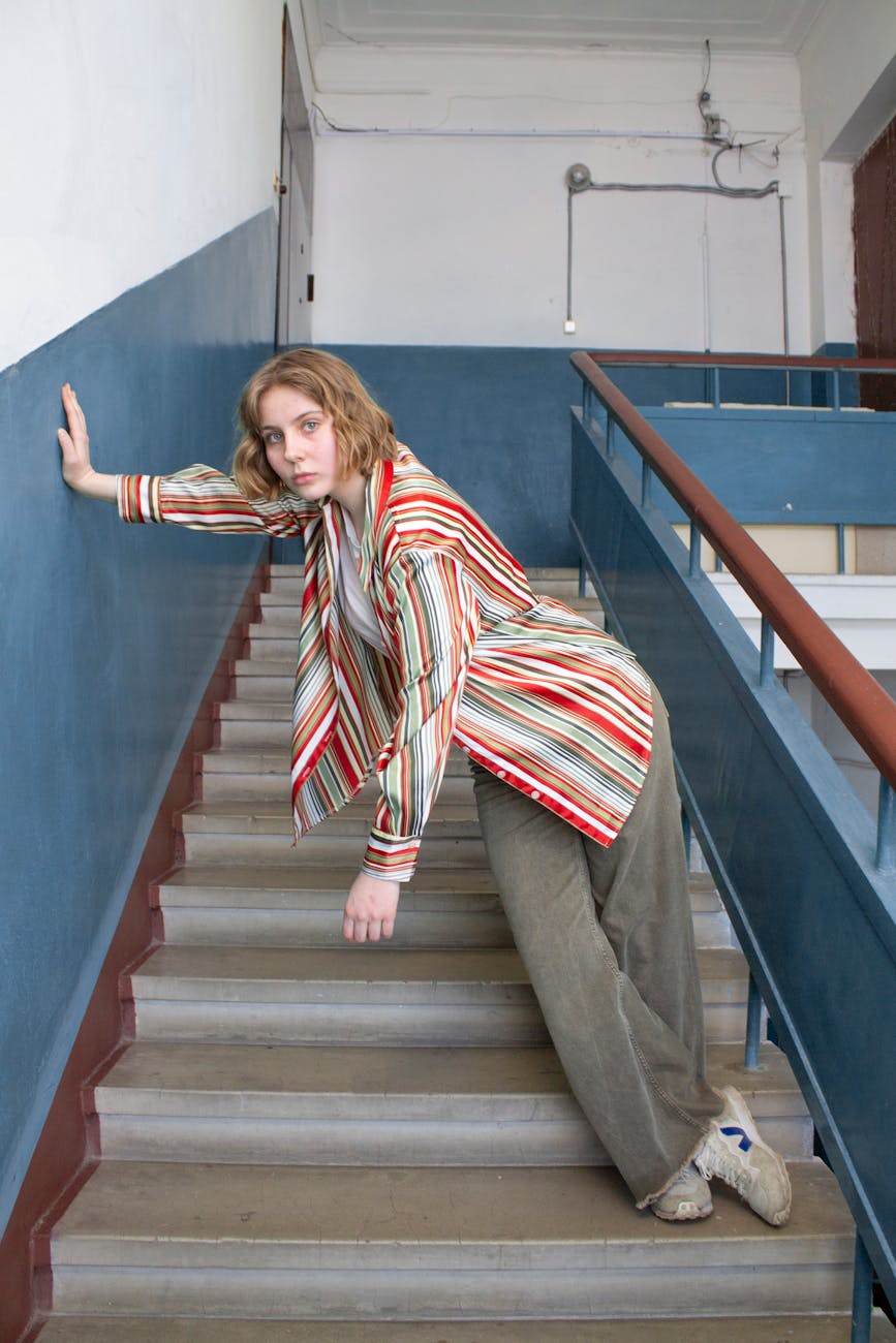 A woman in a red striped shirt stands on stairs while tilting over to one side and holding onto the wall with her hand.