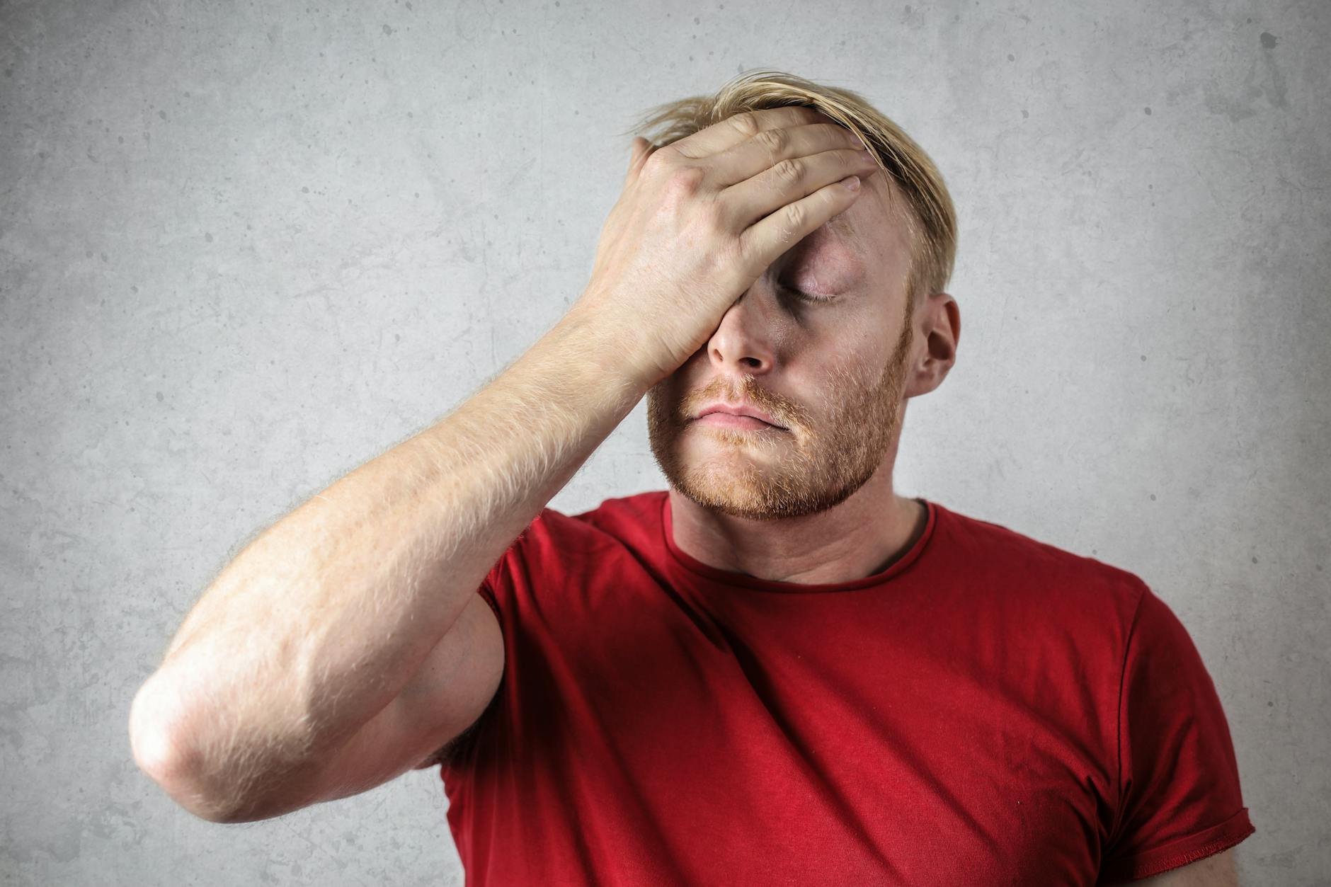 A man with a beard and wearing a red t-shirt holds his hand to his face with his eyes closed.