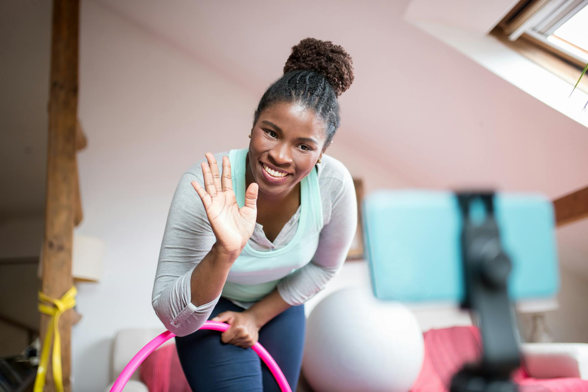 A woman wearing workout clothes holding a hula hoop smiles and waves at the phone camera.