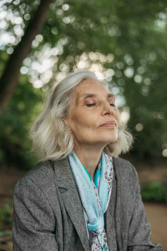 A woman with grey hair wearing a grey coat and blue silk scarf, smiles with her eyes closed and face uplifted.