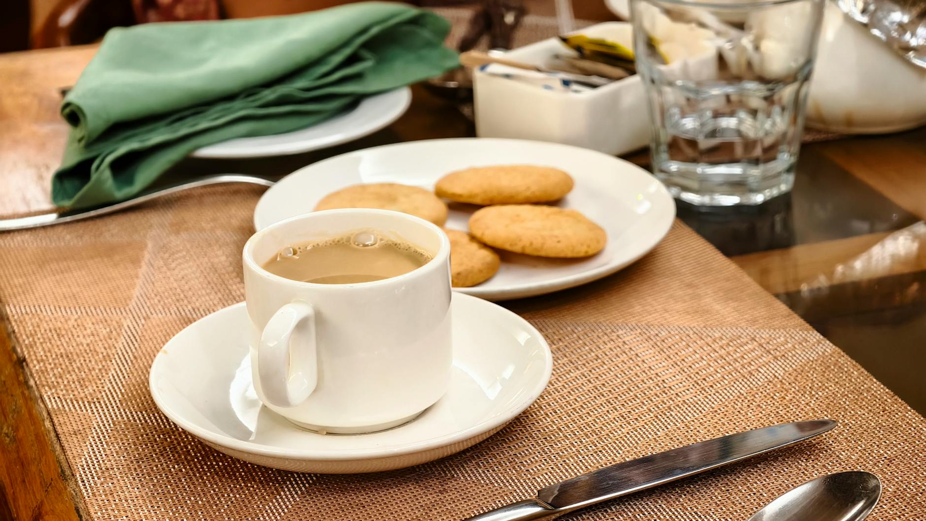 A white cup of milky tea next to a plate of cookies sits on a placemat set for a meal.