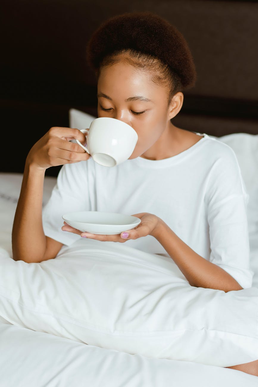 A woman in white sits up in bed and drinks tea from a cup while holding the saucer.