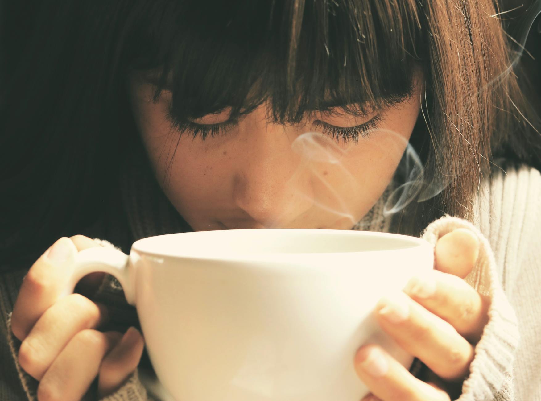 A woman holds a large, white, steaming cup that she's inhaling.