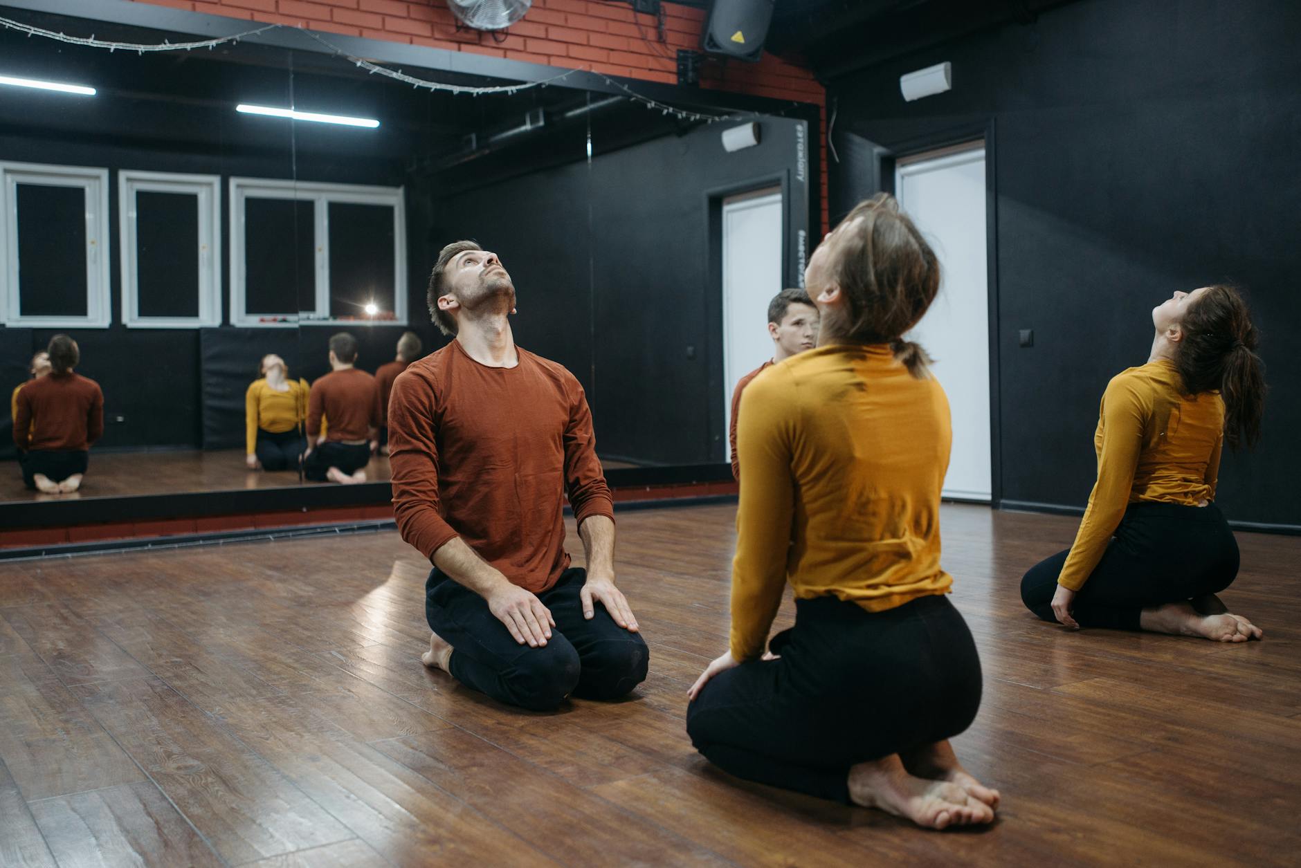 A few people in a doing gentle neck stretches while sitting on their knees in a studio with a mirror on the back wall.