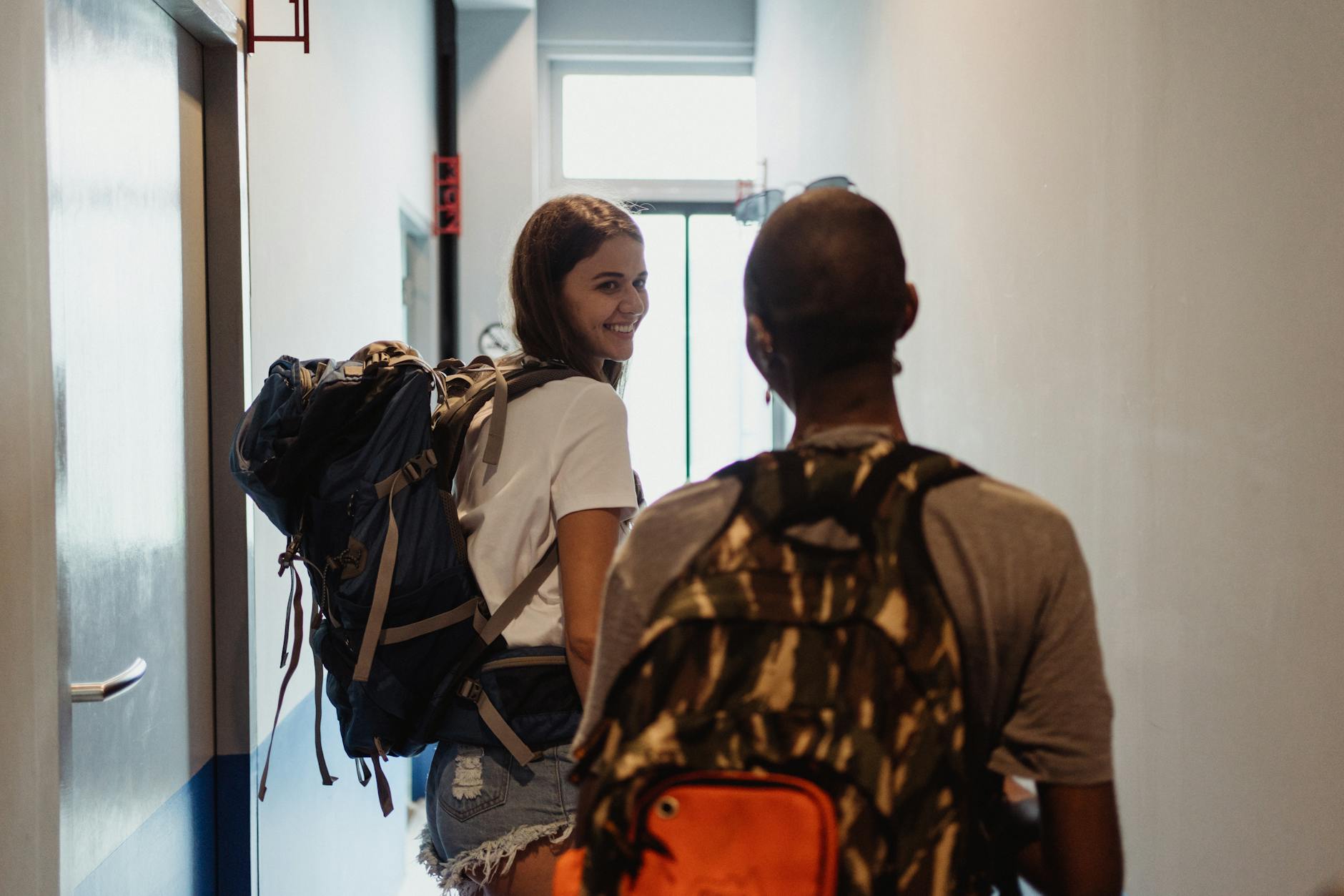 Two young people wearing backpacks walk away down a hall. The first person looks back and smiles at the person behind.