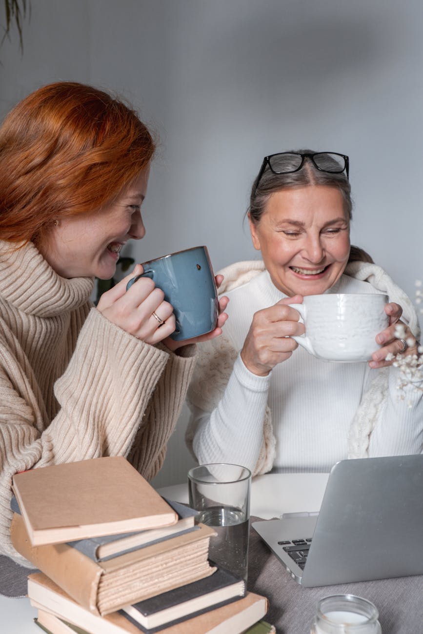Two women smile and hold cups as they look at a laptop. There are books and a glass of water on the table.