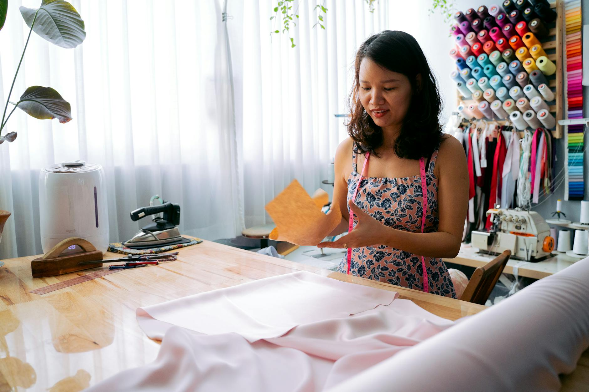 A woman laying out sewing patterns on a table with many colours of thread on the wall behind her.