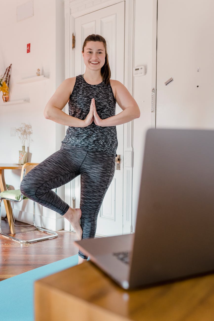 A woman is doing yoga by following an online class on her laptop.