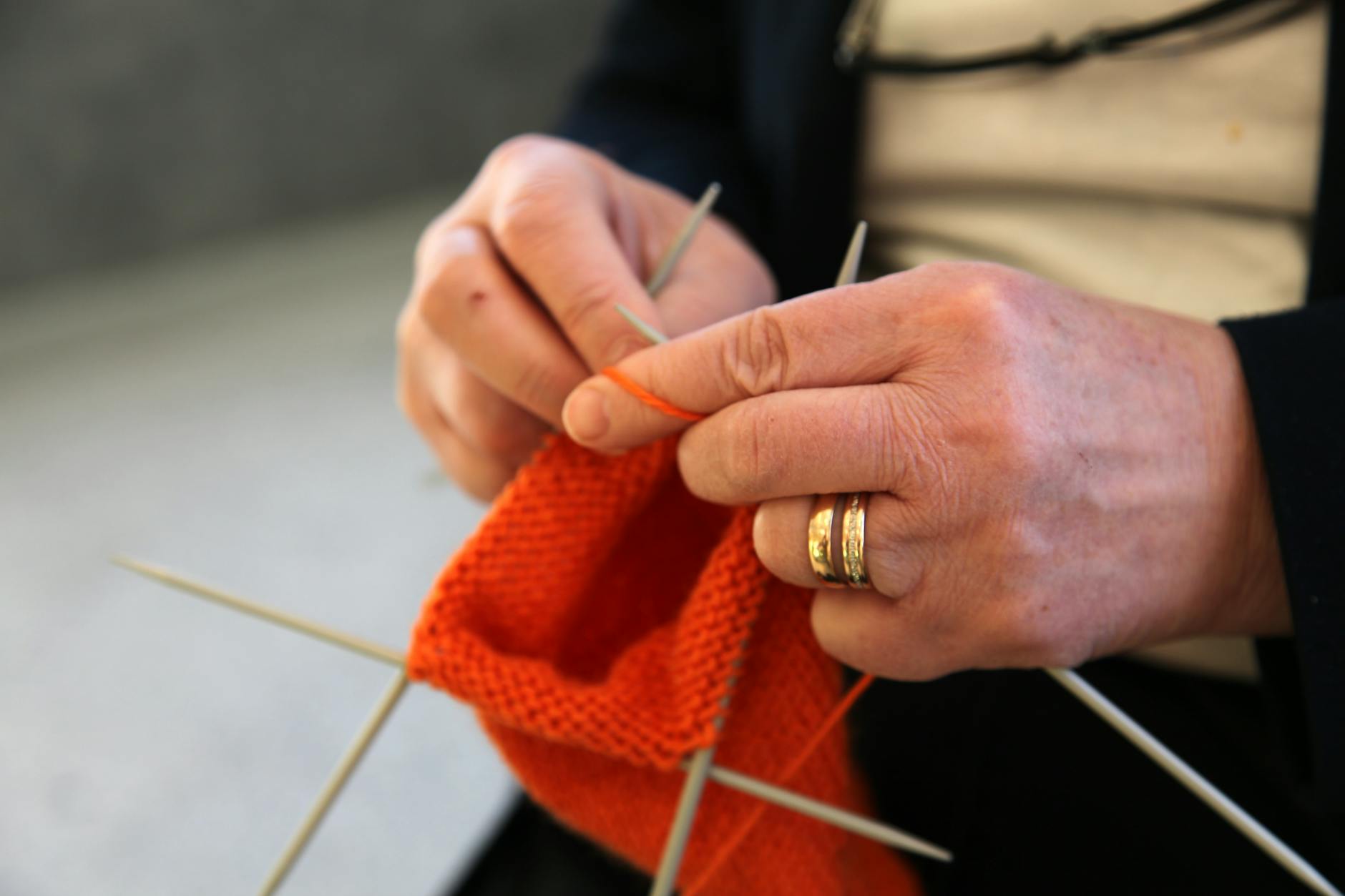 A woman uses four double pointed needles to knit in the round with orange yarn.