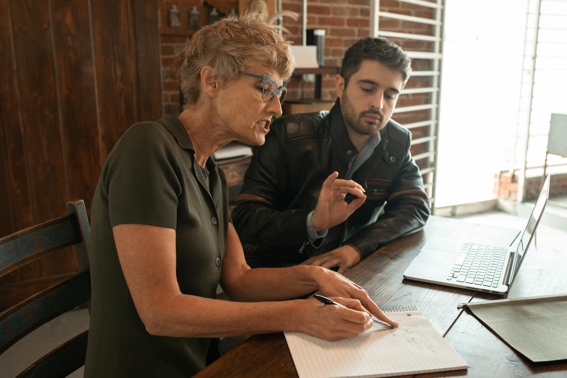 Two people collaborate on ideas while sitting at a table and making notes on a laptop and in a notebook.