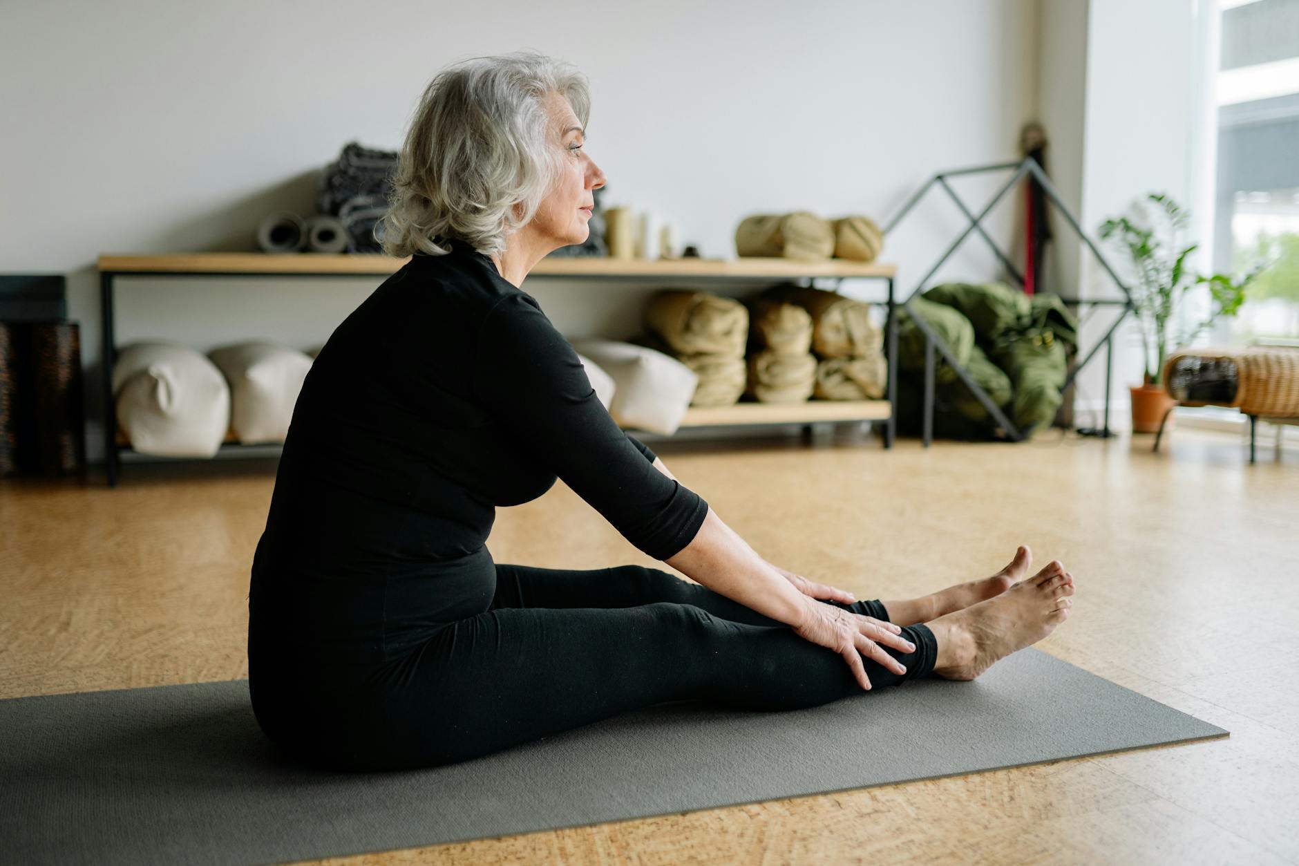 An older woman sits wearing black leggings and a black top sits on a yoga mat in a fitness studio.