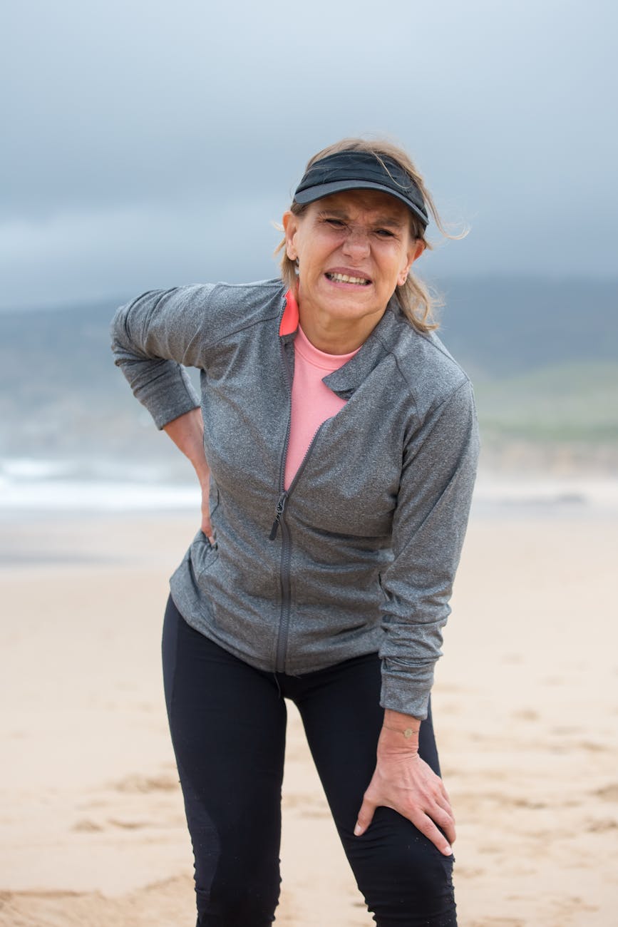 A woman on the beach wearing fitness gear holds her back with one hand and grimaces in pain.