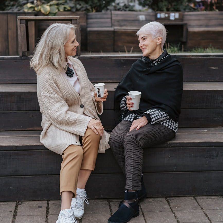 Two women sit on steps holding take-out drinks and chatting.