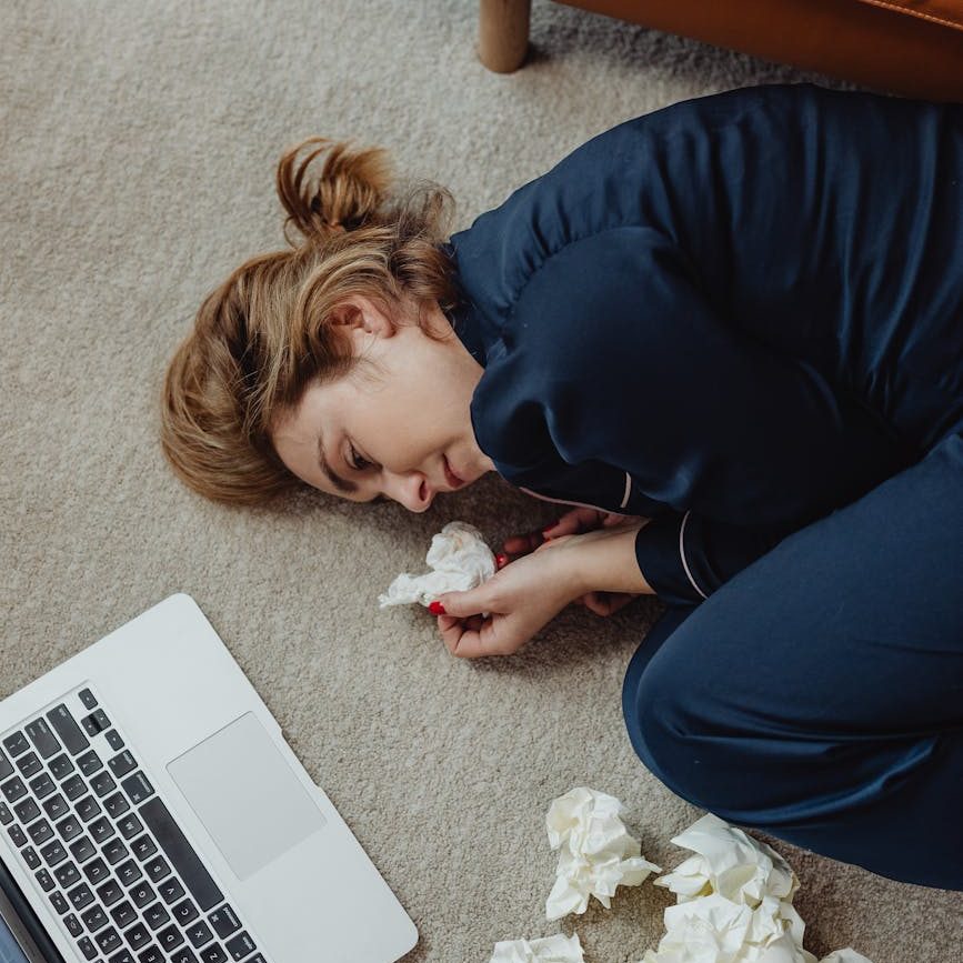 A woman in navy pyjamas curled in a fetal position on the floor next to a laptop and scrunched up papers.