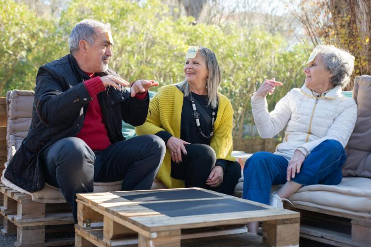 A group of three people sit outside playing a game with paper stuck to their foreheads.