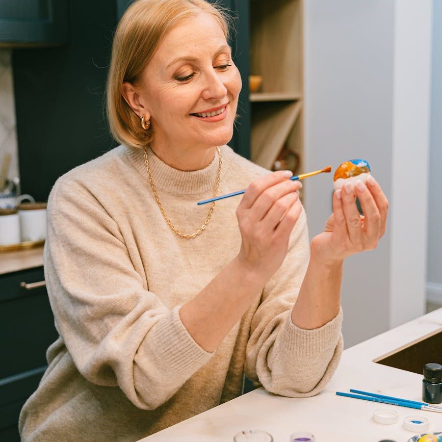 A woman smiling while painting a small object, possibly an Easter egg.