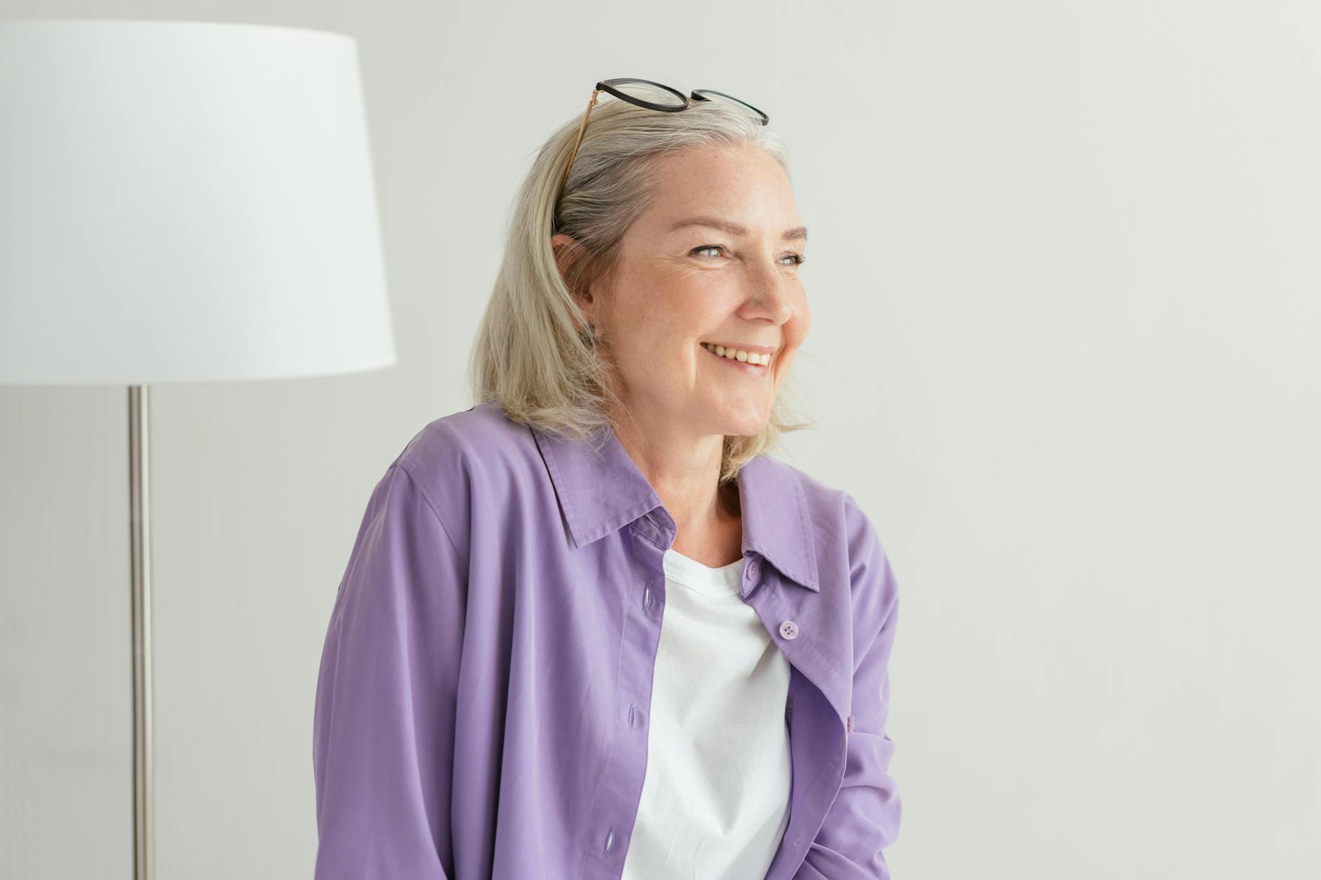 A woman standing next to a lamp with glasses pushed up into her greying hair, smiling off into the distance.