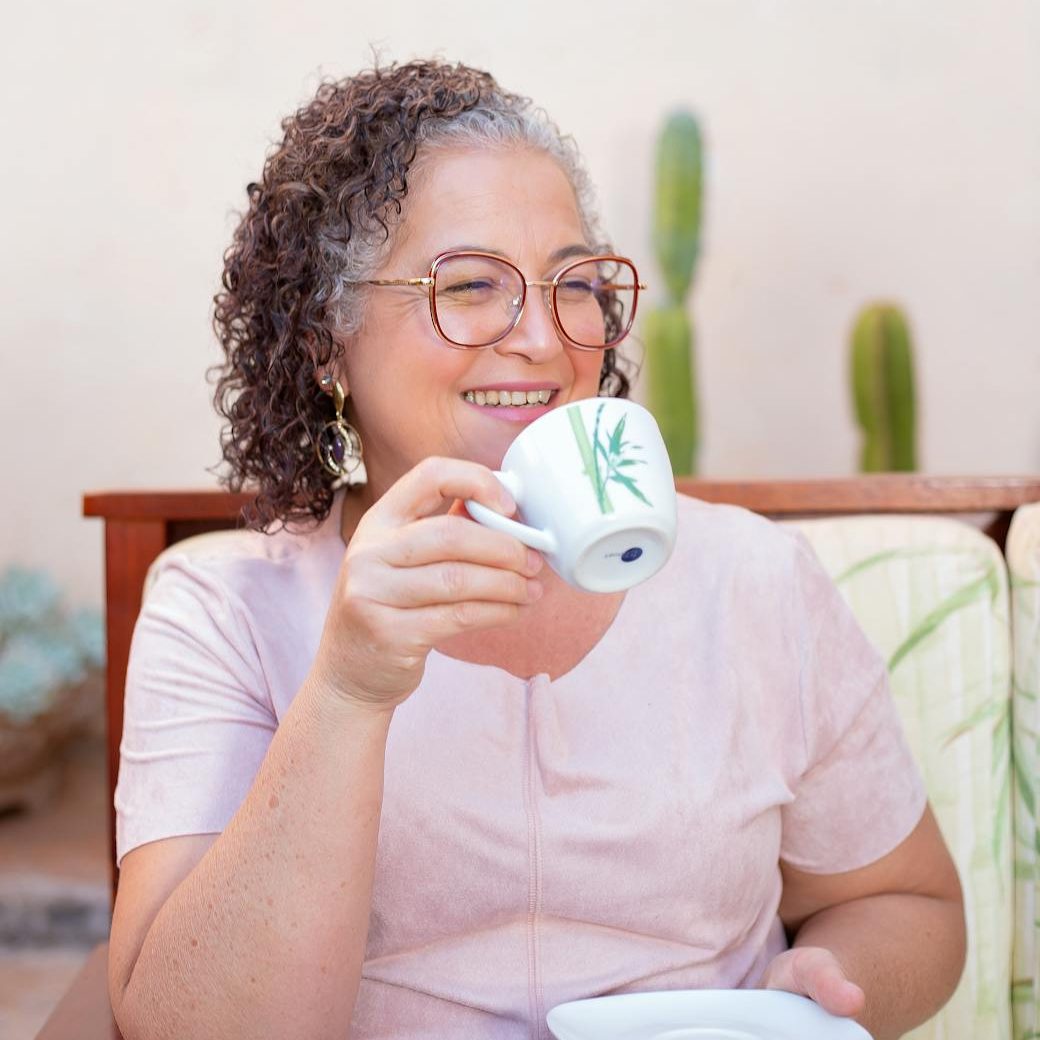 A woman with glasses and greying curly hair smiling as she brings and cup to her mouth.